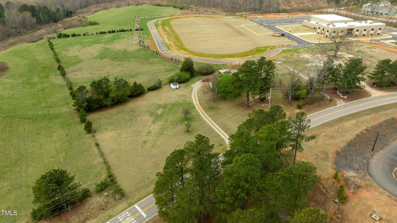 Aerial view of a grassy field with a winding road, trees, a small shed, and a newly constructed building with adjacent parking lot. A sports field is visible near the top of the image.