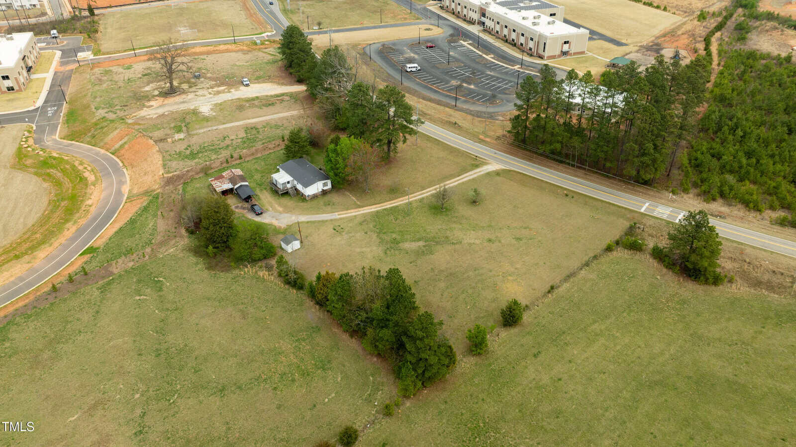 Aerial view of a rural property with a white house, small outbuildings, a curved driveway, and grassy areas. A road runs alongside the property, with commercial buildings and parking lots nearby. Trees border parts of the land.