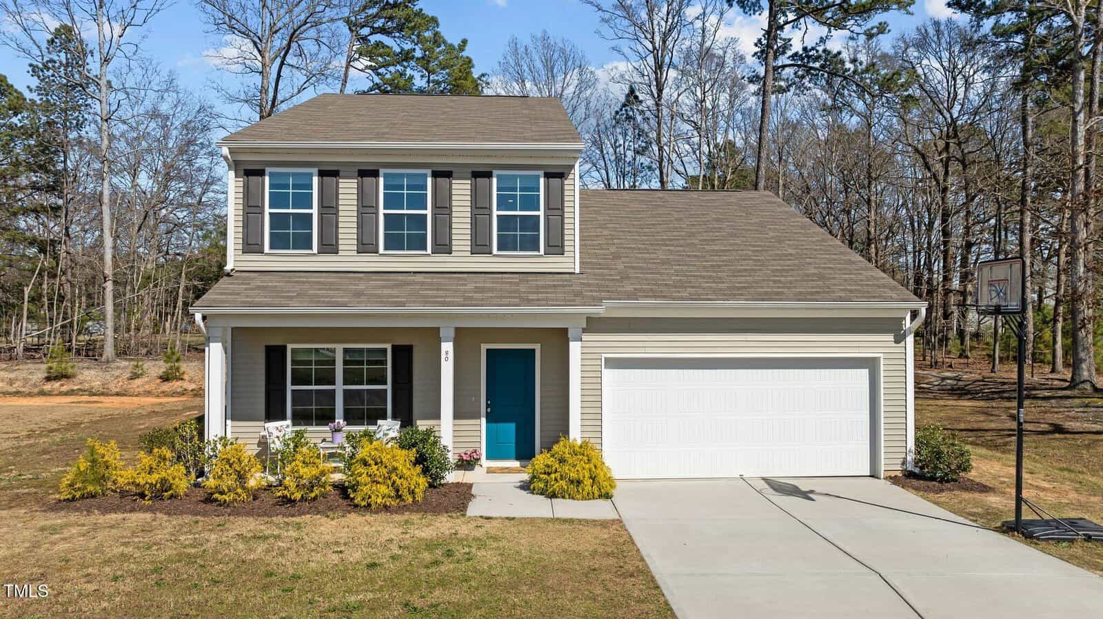 A two-story beige house with white trim, blue front door, and attached two-car garage. There are shrubs along the front and a basketball hoop near the driveway. Trees are visible in the background.