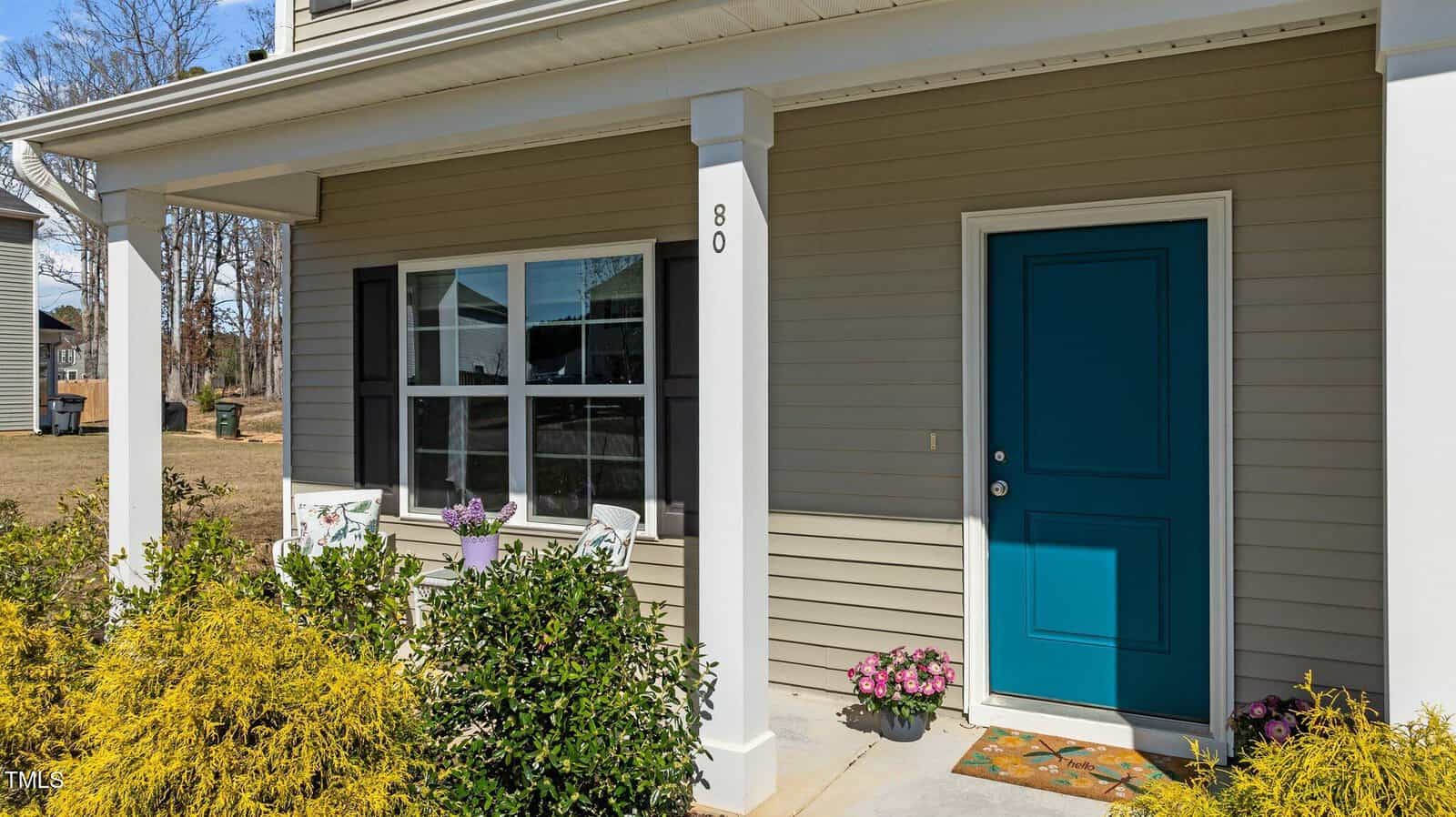 The image shows the front porch of a house with beige siding and a teal door. Two white chairs and a small table sit by the window, surrounded by bushes and potted flowers. The house number "80" is displayed on a white column.