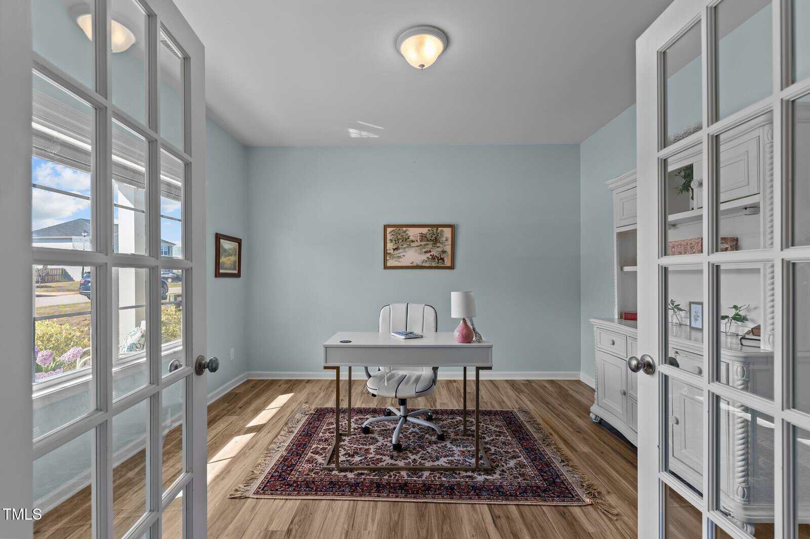 A home office with light blue walls, a desk and chair on a patterned rug, a white cabinet, a table lamp, wall art, and large windows letting in natural light. French doors are partially open in the foreground.