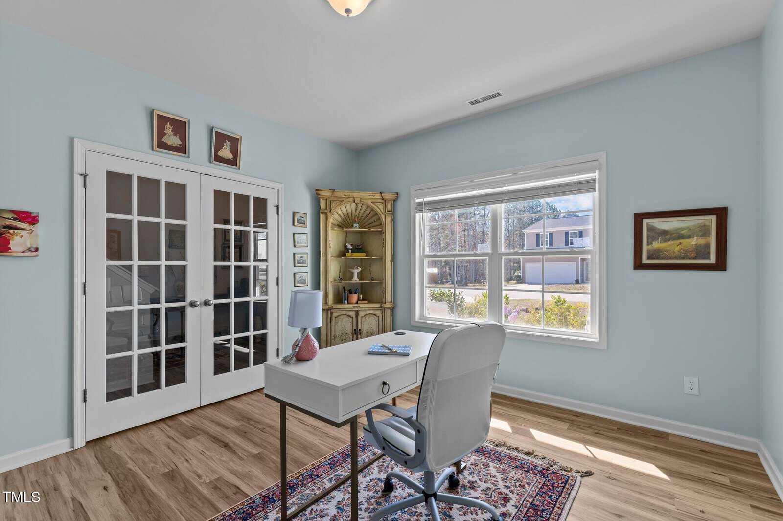 A home office with light blue walls, a white desk and chair, a laptop, a lamp, and books. Sunlight streams in through a large window. Decorative art and framed pictures hang on the walls. Glass double doors are in the background.