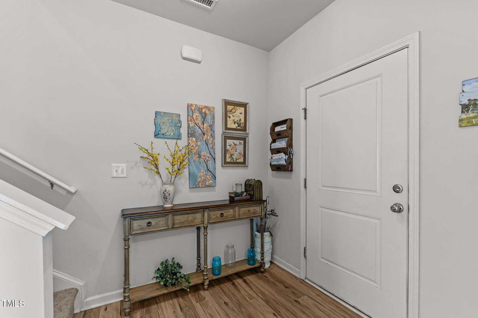 Entryway with a white door, a wooden console table holding a vase with yellow flowers, decor items, and wall art above. Two mail holders are mounted on the wall. Light wood flooring and a staircase are visible.