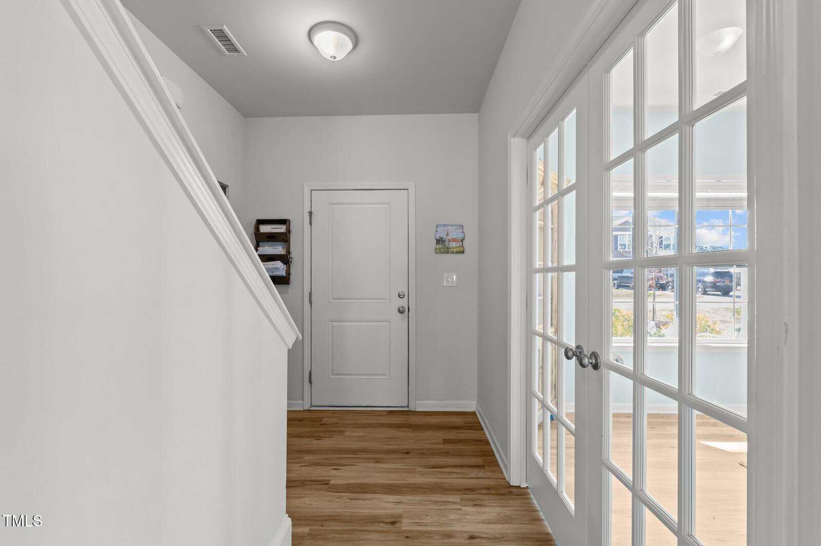 A hallway with light wood floors, white walls, a staircase on the left, a closed white door at the end, wall art, and glass double doors on the right leading to a sunlit room with windows.