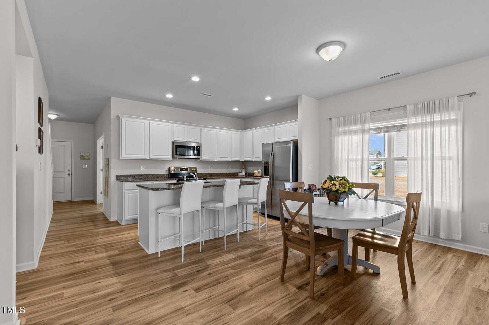 Modern kitchen and dining area with white cabinets, stainless steel appliances, a large island with barstools, and a round dining table with four chairs. Hardwood floors and a window with sheer curtains provide natural light.
