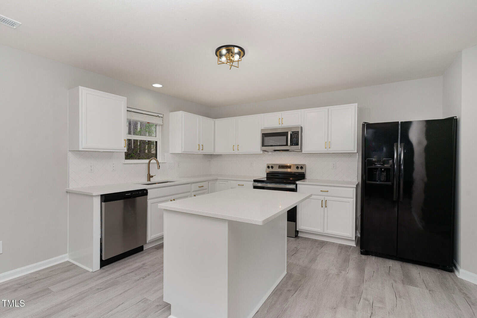 Modern kitchen with white cabinets and a central island. Appliances include a black refrigerator, stainless steel dishwasher, and microwave. The kitchen has wood-patterned flooring, light gray walls, and a window above the sink with a ceiling light fixture.