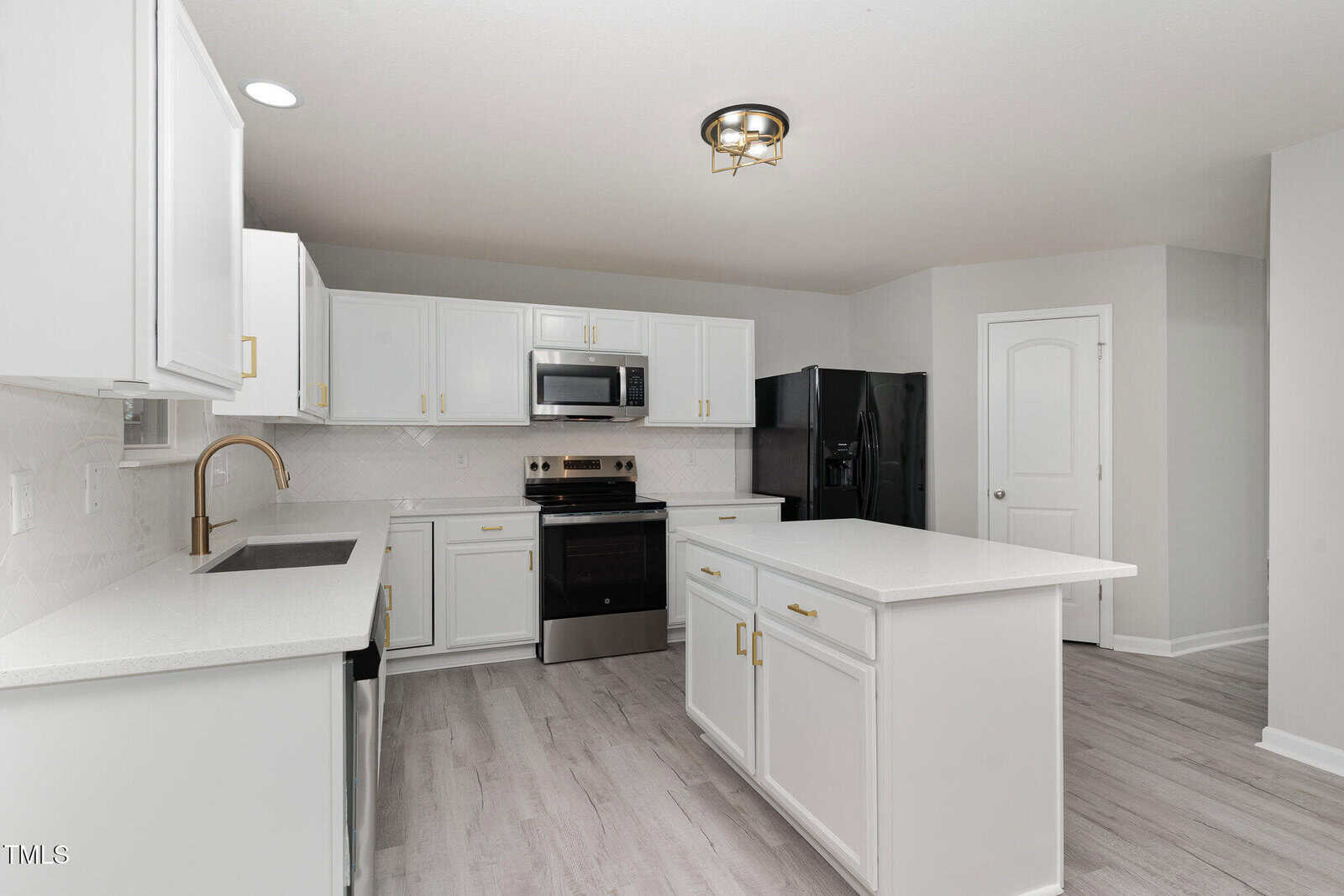 Modern kitchen with white cabinets and countertops, stainless steel appliances, and a center island. Light wood flooring contrasts with dark refrigerator. Recessed lighting overhead and a single door on the right wall.