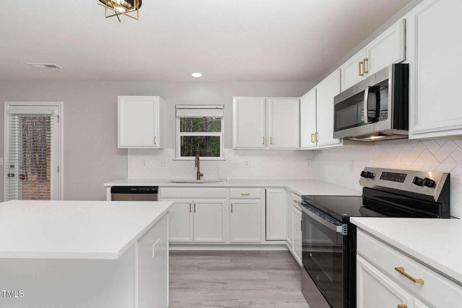 Modern kitchen with white cabinets, a central island, and stainless steel appliances including a stove and microwave. There is a window above the sink, which has a view of trees outside. The floor is light-colored wood, and the walls are neutral.