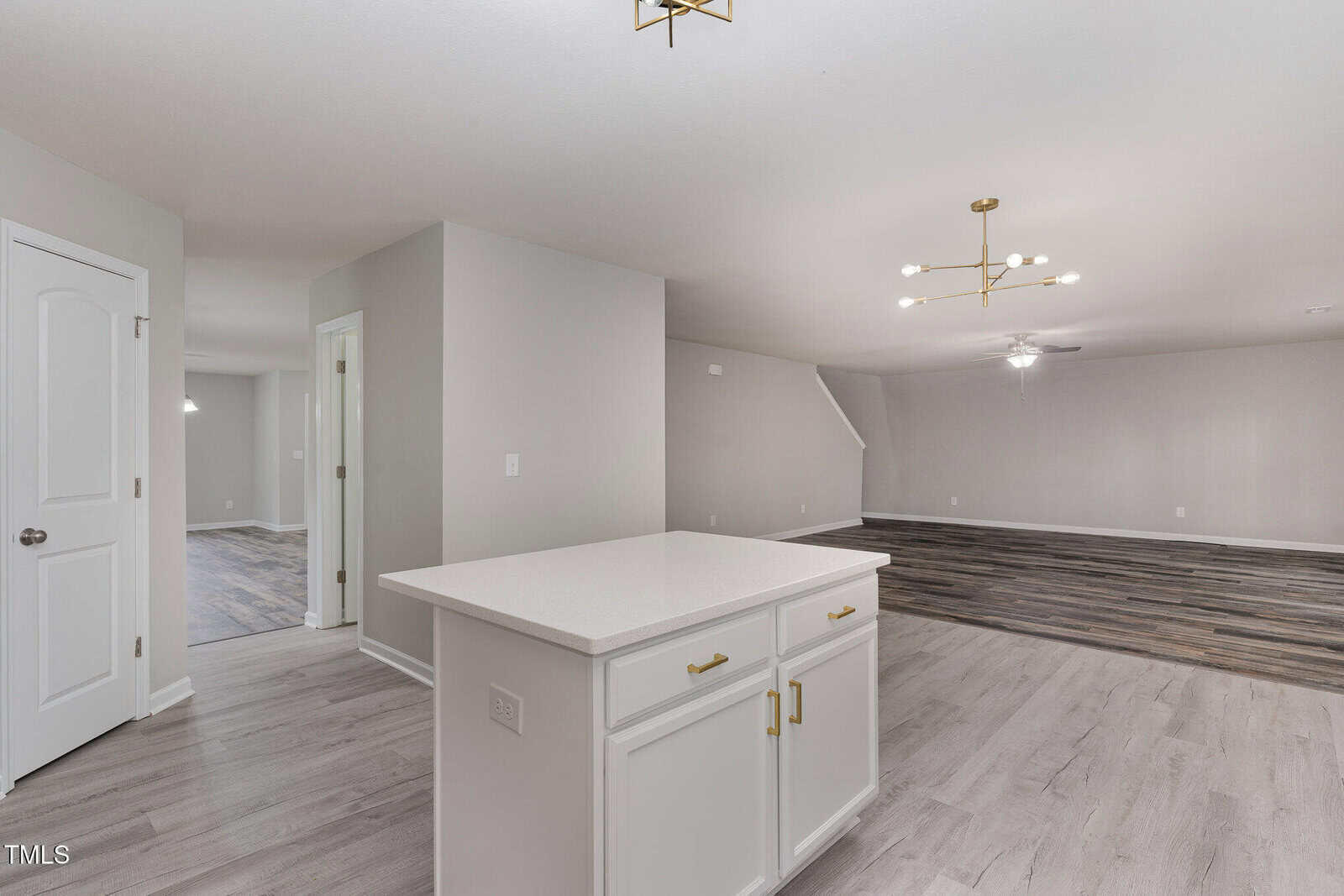 A modern, open-concept interior features a kitchen area with a white island and cabinets. The space opens into a living room with wood-like flooring, a staircase, and contemporary light fixtures. Walls and ceiling are painted light gray.