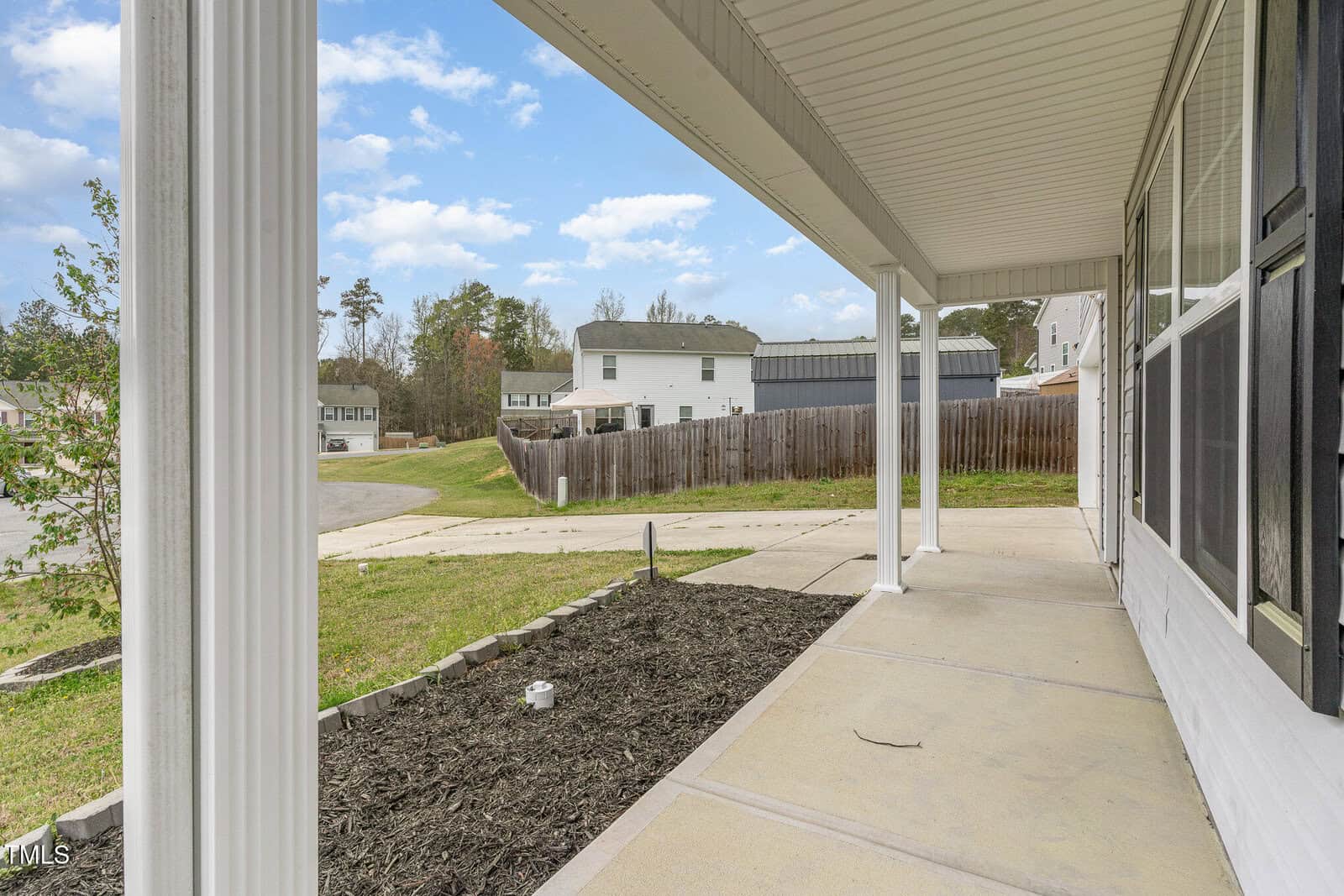 View from a covered front porch overlooking a residential neighborhood. The porch features white columns, a black railing, and a concrete floor. A mulched garden bed is on the left, and a curved street with houses is in the background.