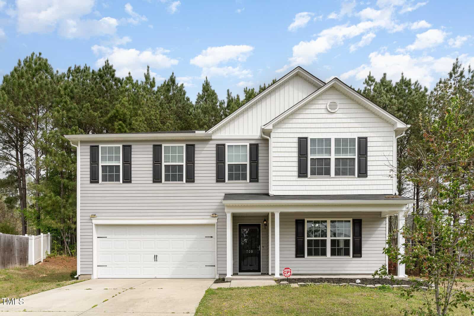 A two-story house with white siding and black shutters is shown. It has a two-car garage and a small front porch. The driveway is concrete, and the background features trees and a partly cloudy sky.