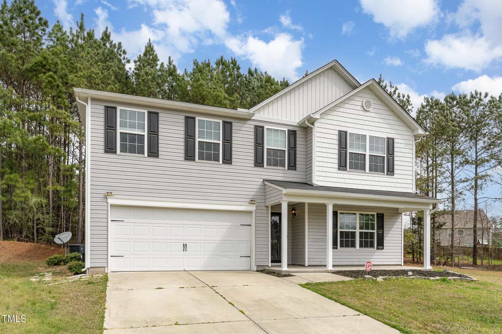 A two-story house with light gray siding and black shutters. It features a double garage door, several windows, and a front porch with columns. The driveway leads to a small lawn area. Trees are in the background under a cloudy sky.