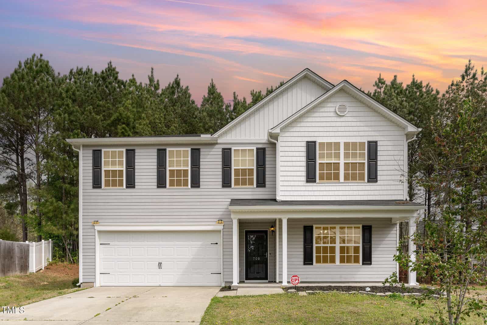 The two-story white house with black shutters and a two-car garage sits gracefully at 709 Conifer Court in Creedmoor, surrounded by lush greenery. A colorful sunset graces the sky, while a small tree and paved driveway add charm to the picturesque setting.