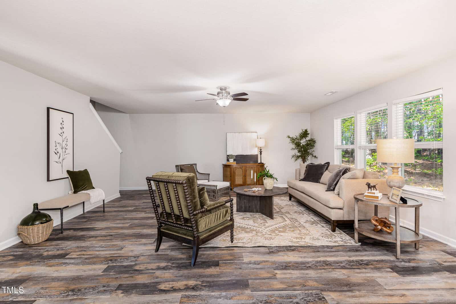 A spacious living room with wood flooring, a beige sofa, two armchairs, a round coffee table, a patterned rug, and large windows letting in natural light. There is a bench and a potted plant near the staircase.