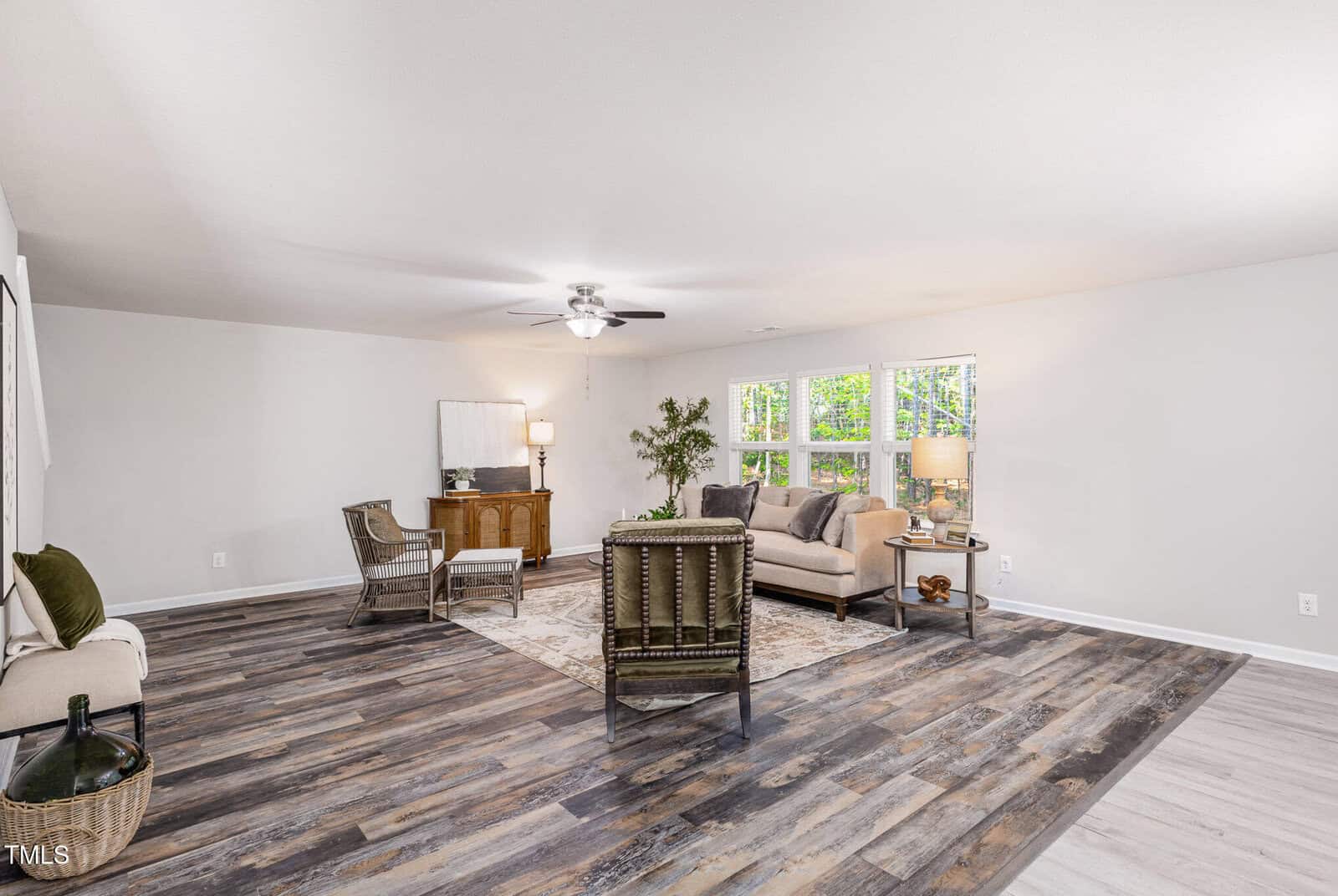 Spacious living room with wood-look flooring, neutral walls, and a ceiling fan. Room includes a sofa, armchairs, side tables, and large windows letting in natural light. A desk and plant are placed near the wall.