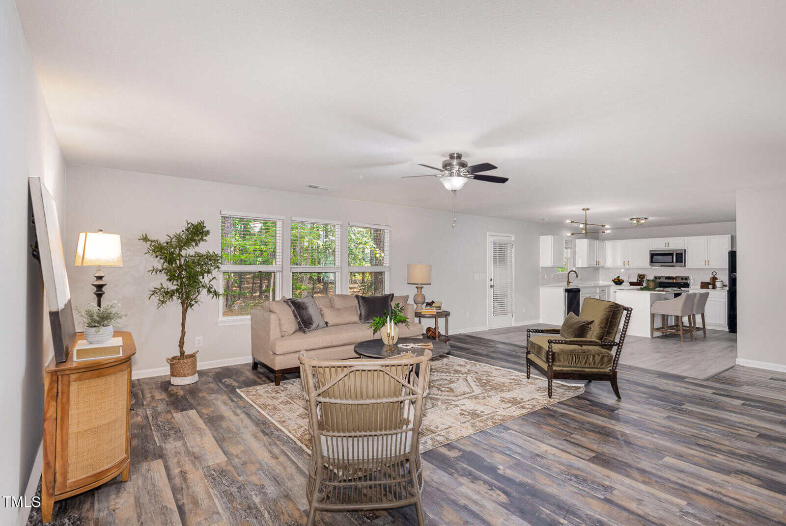 A spacious open-concept living area with wood-style flooring, neutral walls, a sofa, chairs, and rug. Large windows let in natural light. The kitchen with white cabinets and an island is visible in the background.