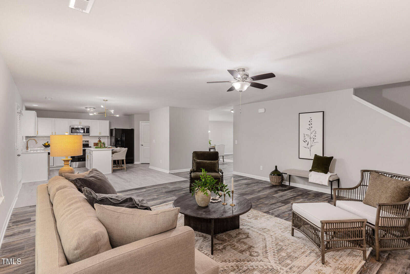 A spacious living room with a beige sofa, round coffee table, and two chairs next to a staircase. The open layout connects to a dining area and kitchen. Neutral colors and minimalist decor are featured throughout.