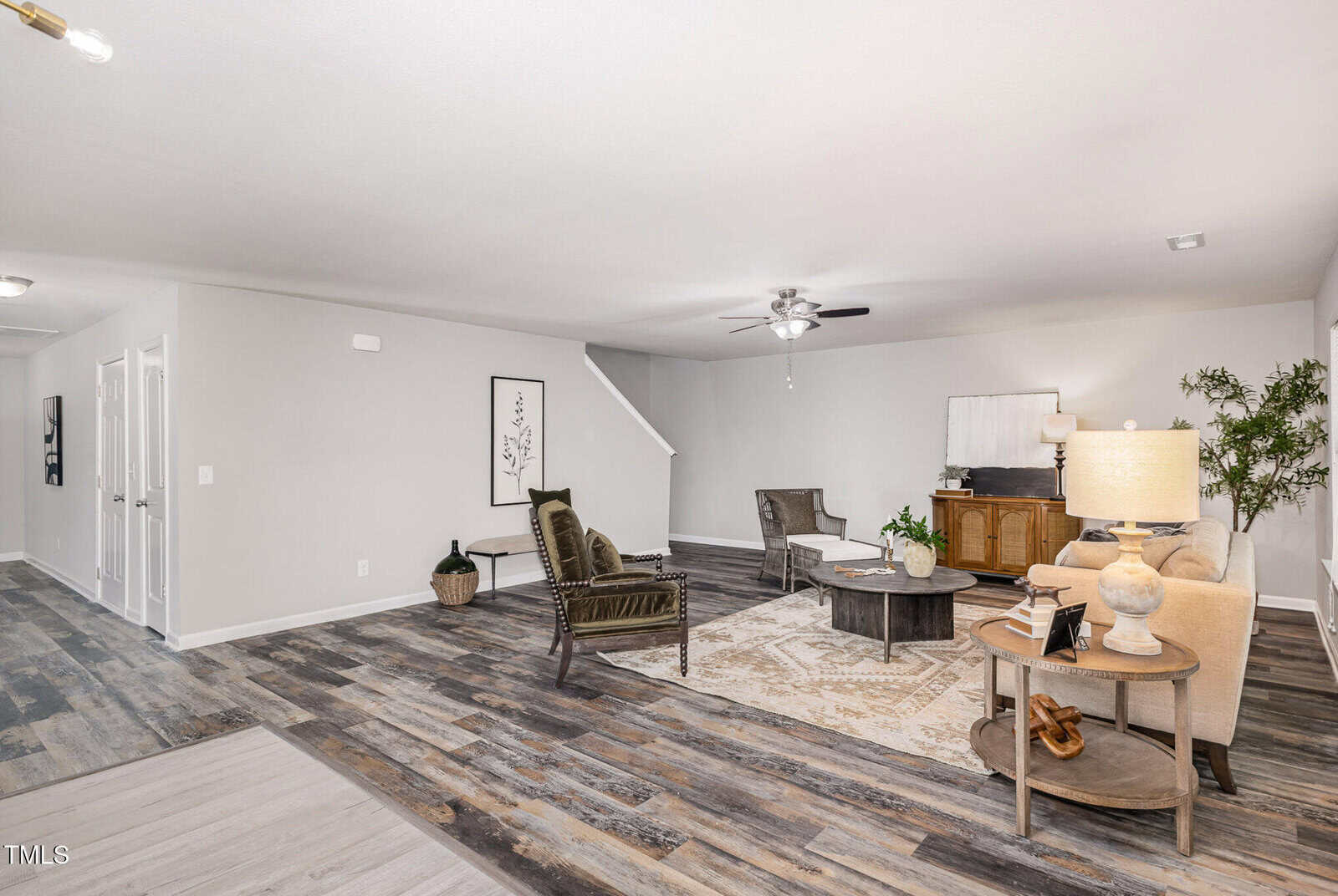 A spacious living room with wood-look flooring, neutral walls, and a ceiling fan. The room is furnished with a beige sofa, two dark chairs, a wooden cabinet, two round coffee tables, and a large area rug.