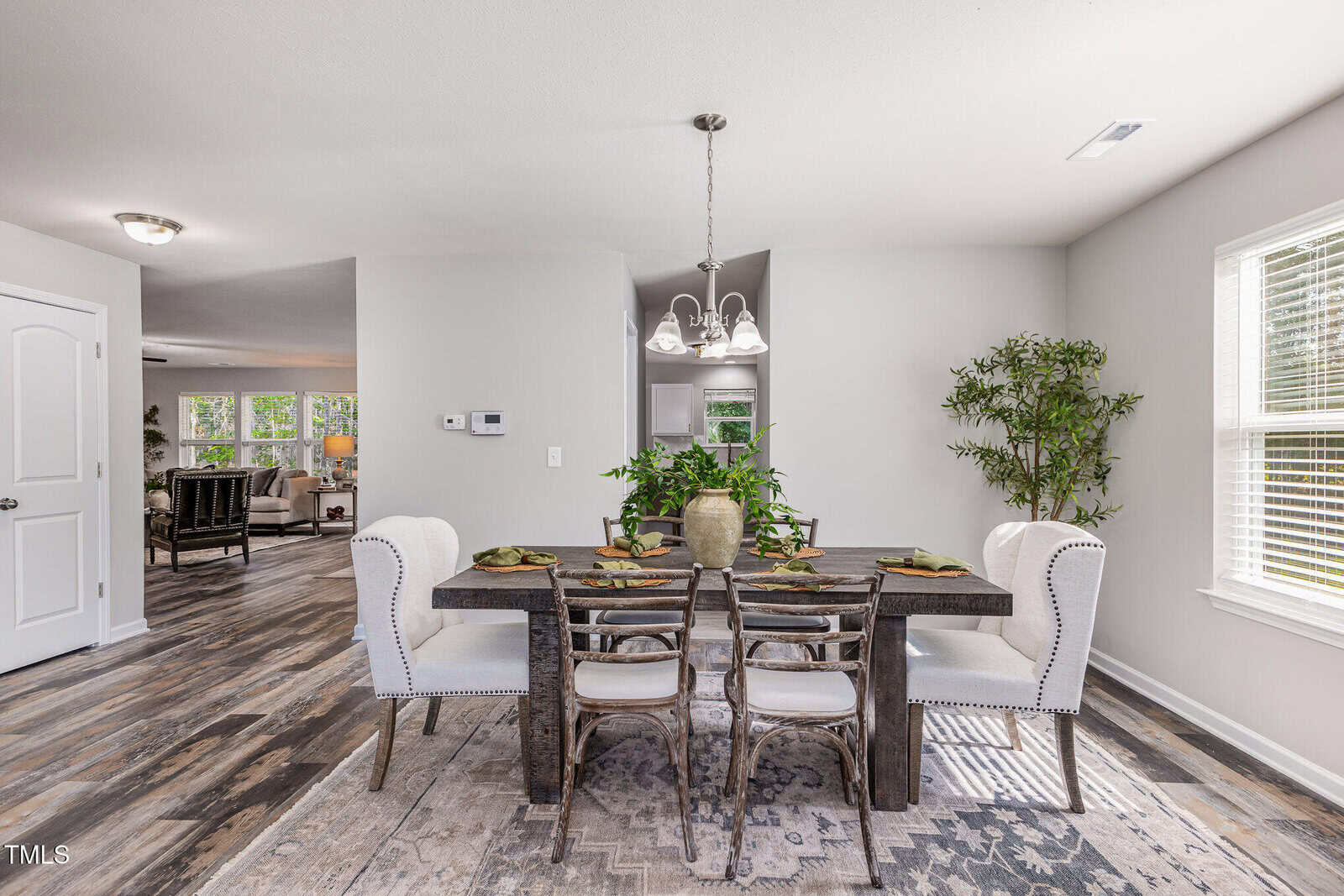 A dining room with a dark wooden table set for six, surrounded by white upholstered chairs. The floor is wood, walls are light gray, and two potted plants sit by the window. A modern chandelier hangs above the table.