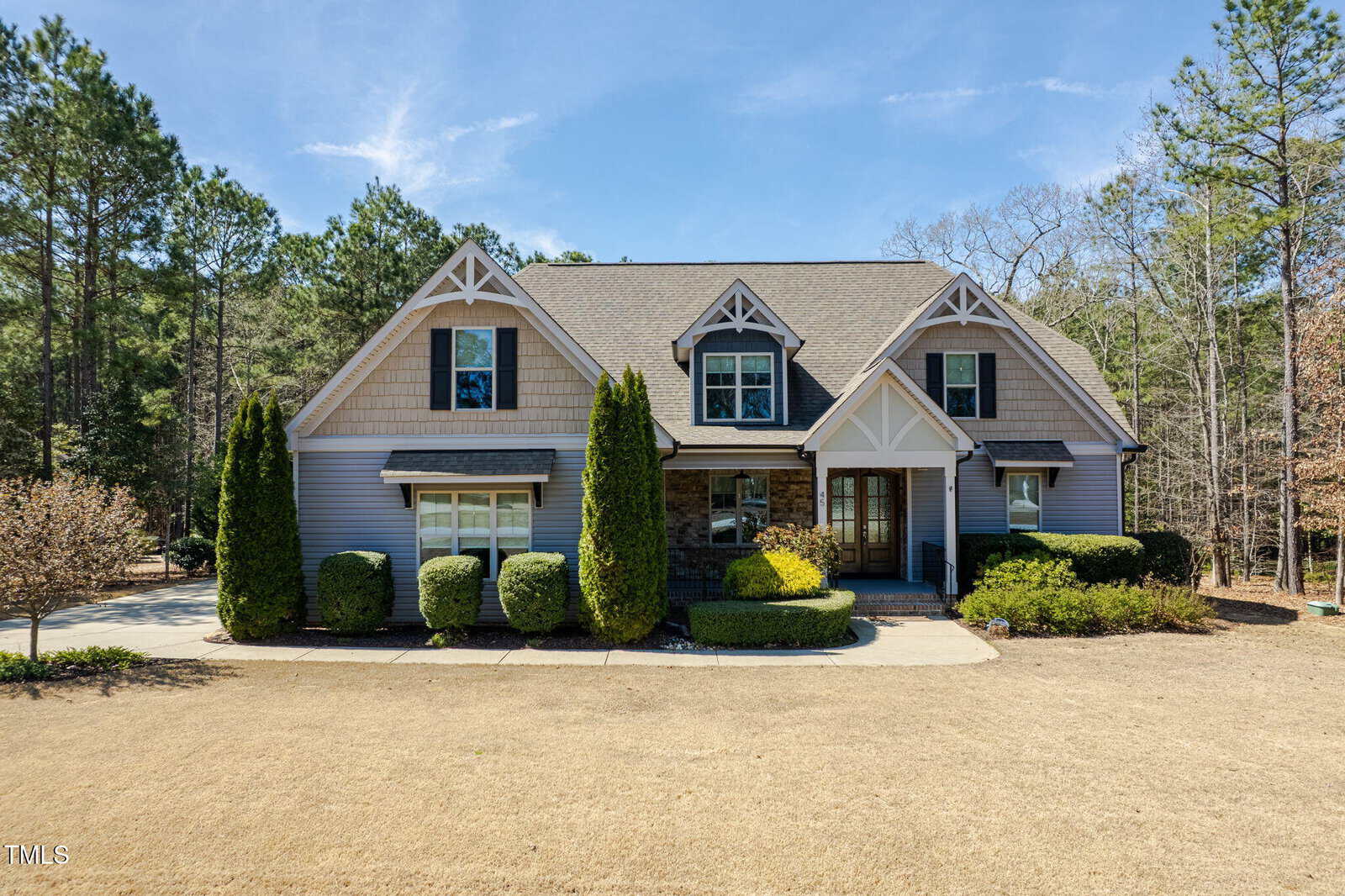 A two-story suburban house with gray siding, stone accents, and a gable roof, surrounded by neatly trimmed bushes and tall trees, with a dry, brown lawn in front.