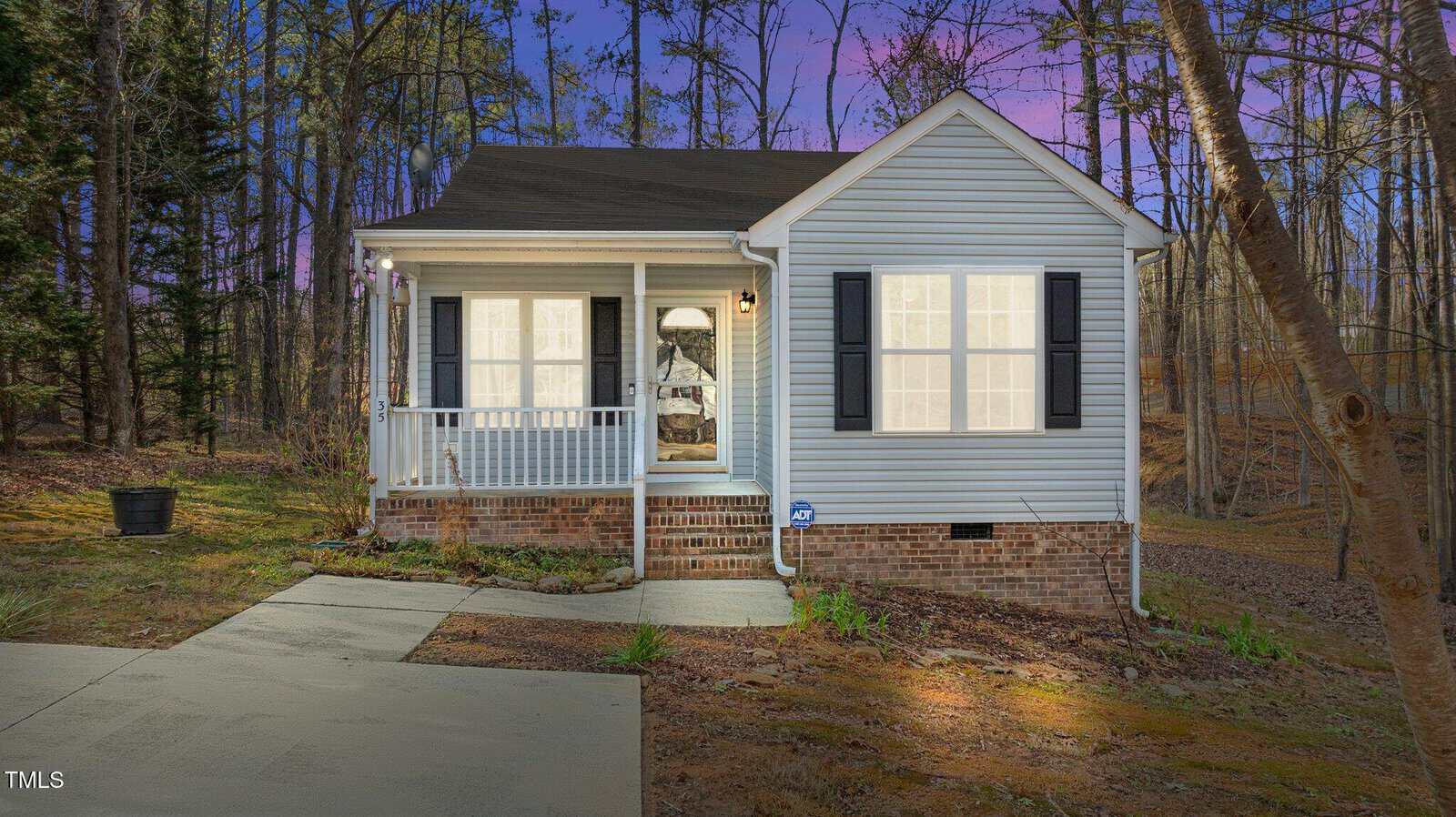A small, single-story house with light gray siding, black shutters, and a front porch with white railings sits on a brick foundation. The house is surrounded by trees and a driveway is visible in the foreground.