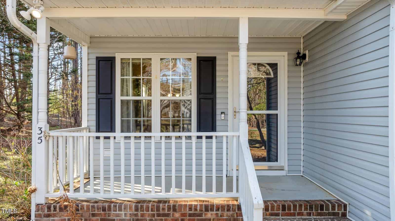 Small front porch with white railing and columns, brick steps, a glass storm door, a window with black shutters, and light gray siding. The house number 35 is visible on a porch post. Trees can be seen in the background.