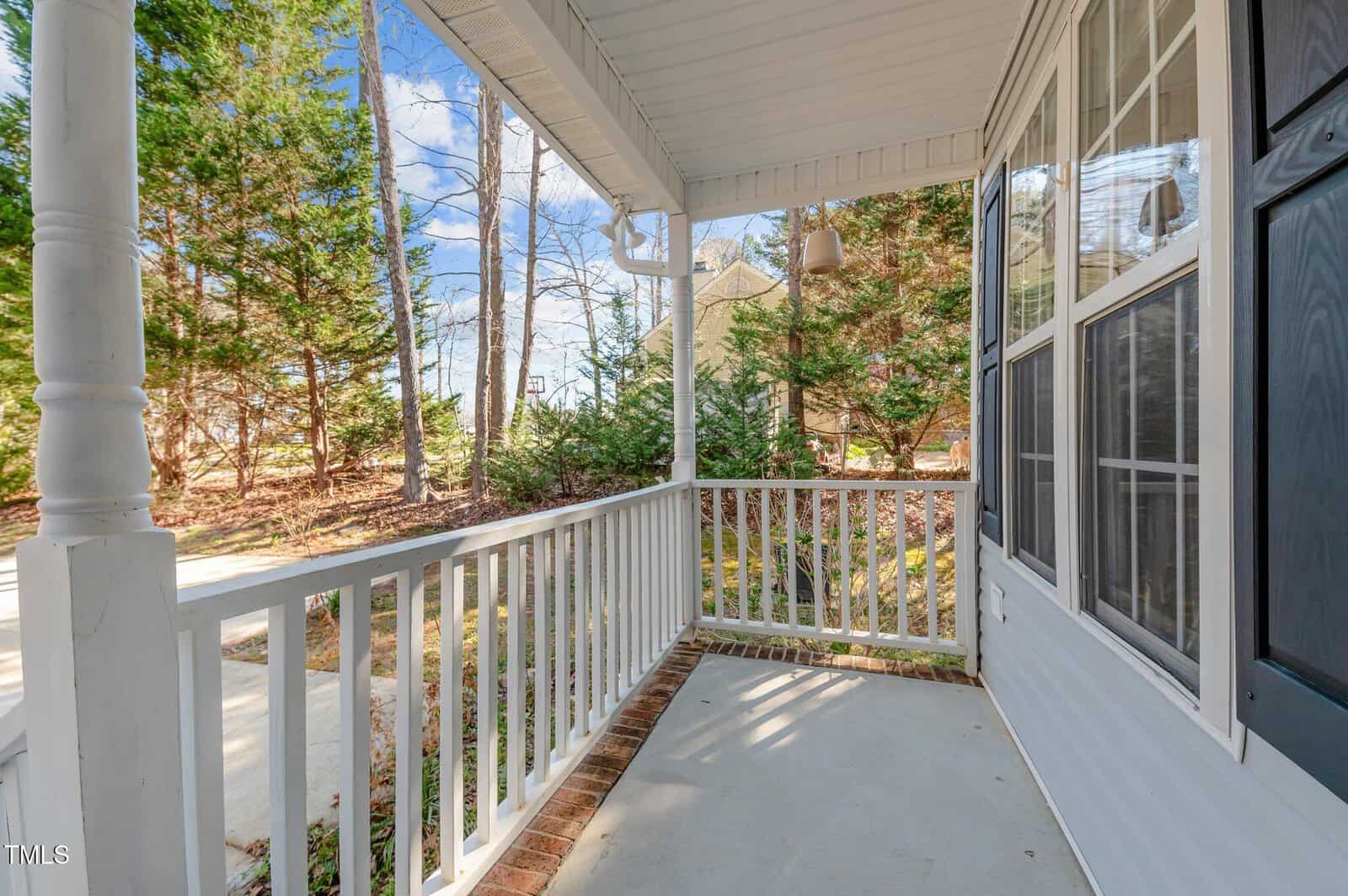 A covered front porch with white railing and a concrete floor attached to a house with white siding and large windows, overlooking a driveway and surrounded by trees.