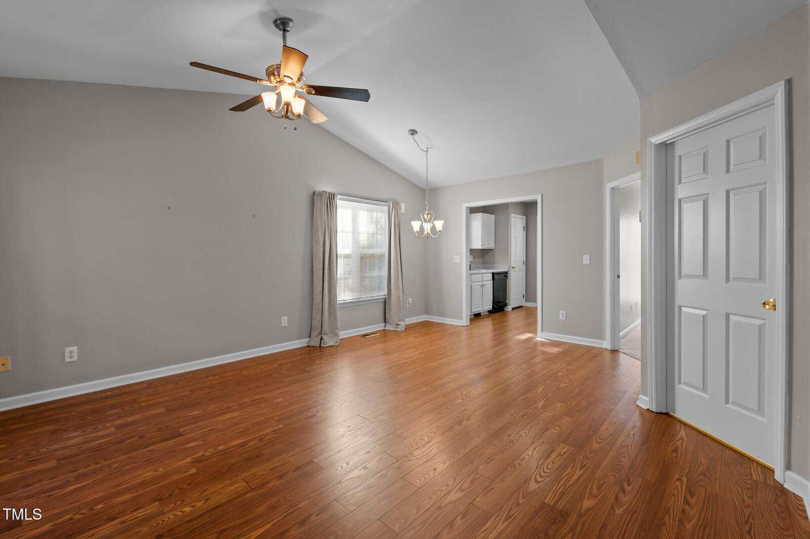 Unfurnished living room with wood flooring, gray walls, and a ceiling fan with lights. Two windows with curtains, an open doorway to a kitchen, and a chandelier hanging over the dining area are visible.