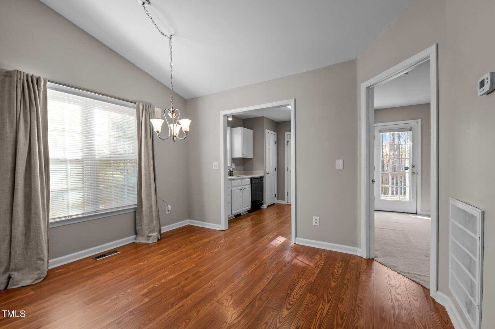 Dining area with hardwood floors, beige walls, a window with curtains, chandelier, and view into a kitchen with white cabinets and an adjacent carpeted room with a glass door leading outside.