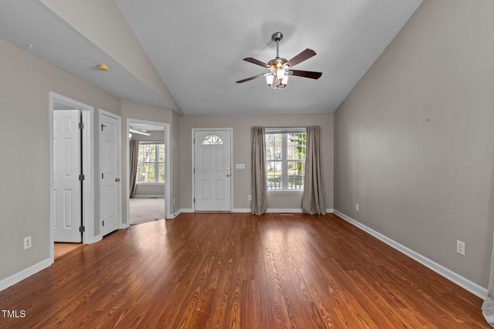 Empty living room with wood floors, gray walls, ceiling fan with lights, a front door, window with beige curtains, and open doorways leading to other rooms. Natural light comes in from the window and door.