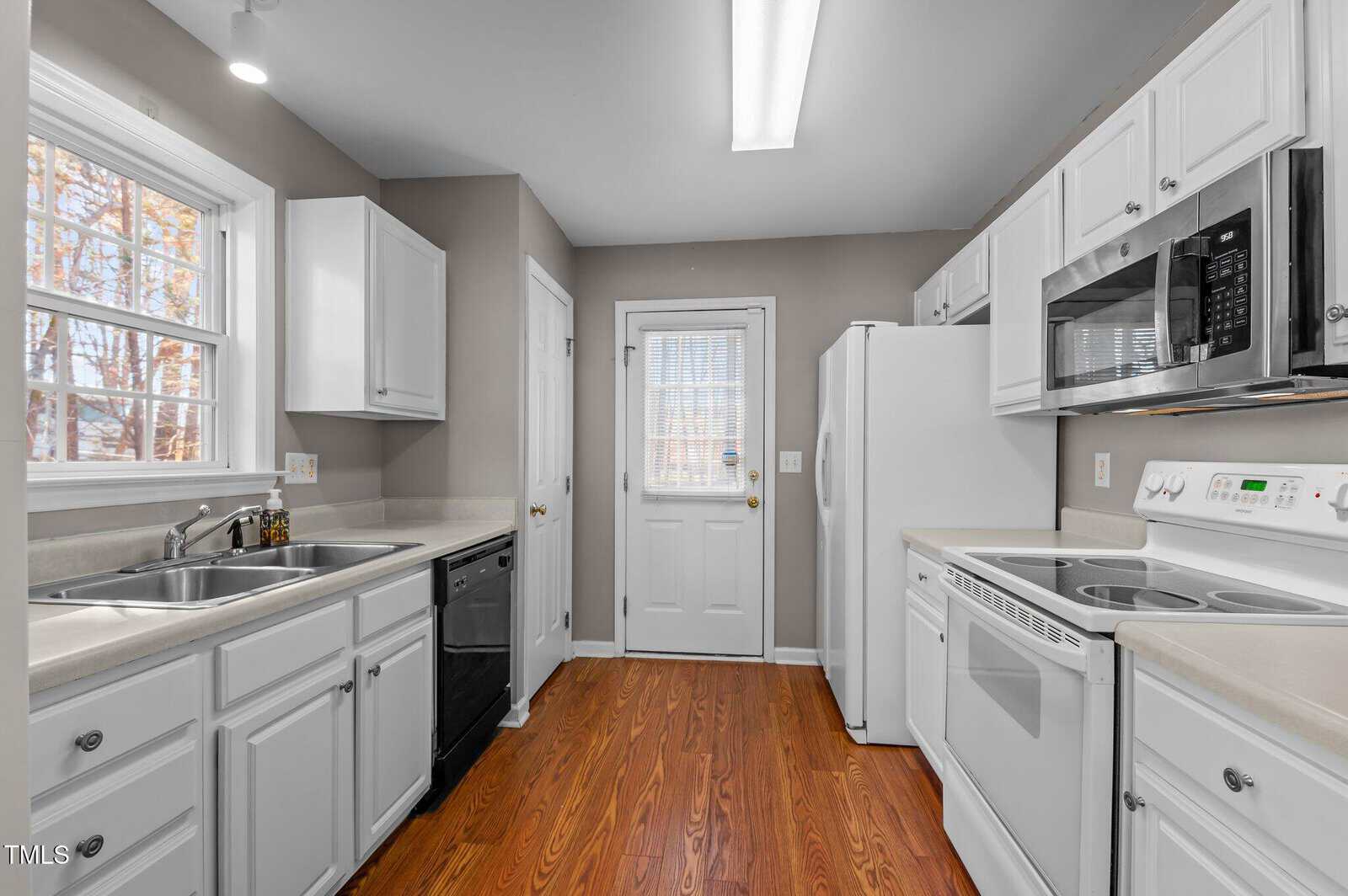 A kitchen with white cabinets, white appliances, a double sink, a window above the sink, a dishwasher, wood floors, and a door with a window leading outside. Neutral-colored walls and ceiling lighting are visible.