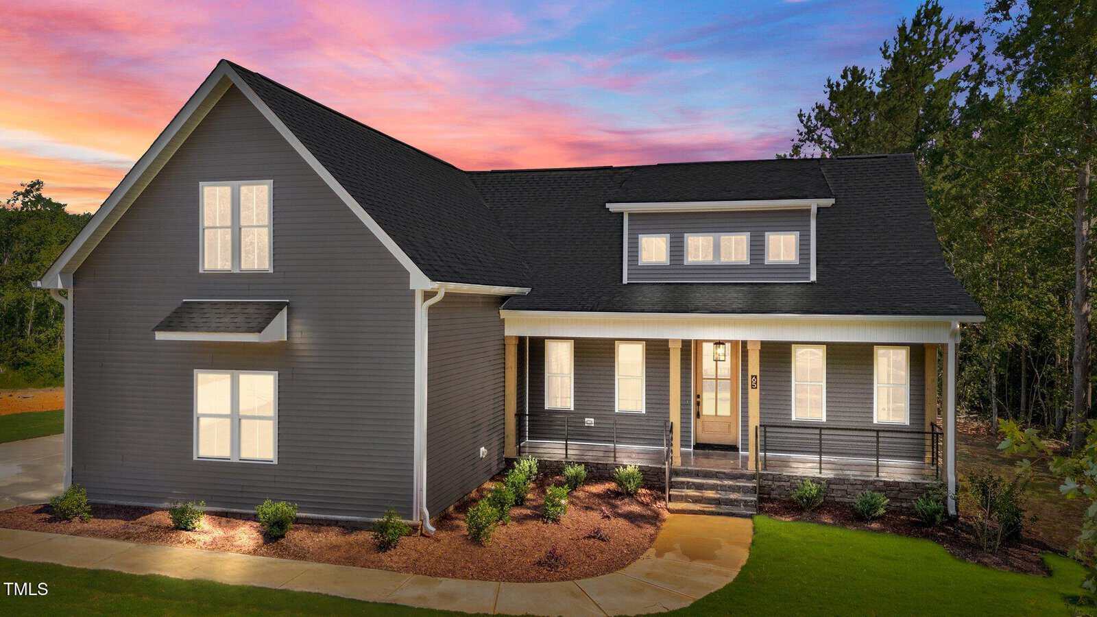 A modern gray house with a large front porch, white trim, and multiple windows, surrounded by landscaping and trees. The sky is vibrant with shades of pink, orange, and purple at sunset.