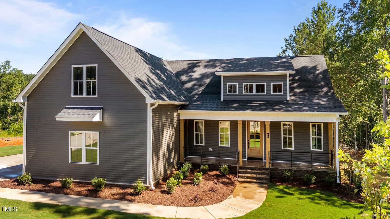 A modern gray house with white trim at 65 Stream View Way, Zebulon, features large windows and a covered front porch. Landscaped plants and trees surround the home, while a concrete path leads to the inviting front door.