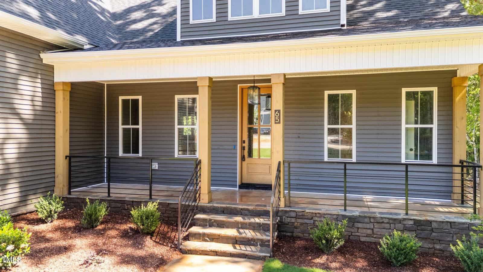 A modern house front with gray siding, a wooden front door, large windows, a covered porch with wooden pillars, black metal railings, stone steps, and neatly landscaped bushes.