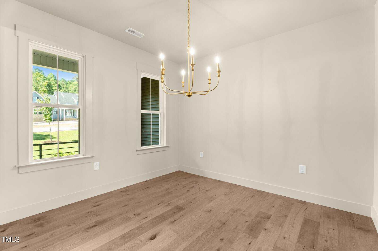 A bright, empty room with light wood floors, cream-colored walls, a gold chandelier, and two windows letting in natural light. A suburban house and greenery are visible outside the windows.