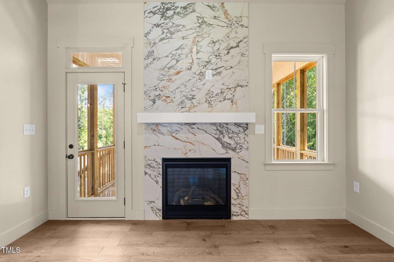 A modern living room features a marble accent wall with a built-in fireplace, light wood floors, a glass door to a porch, and a large window, creating a bright and elegant space.