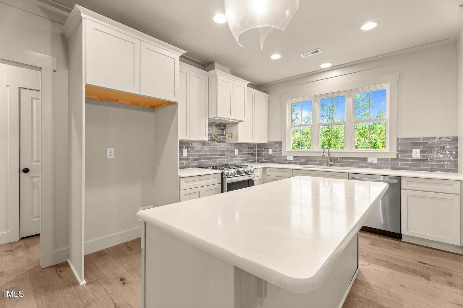 Modern kitchen with white cabinets, a large central island, light wood floors, stainless steel appliances, gray tile backsplash, and three windows above the sink letting in natural light. A space for a refrigerator is visible.