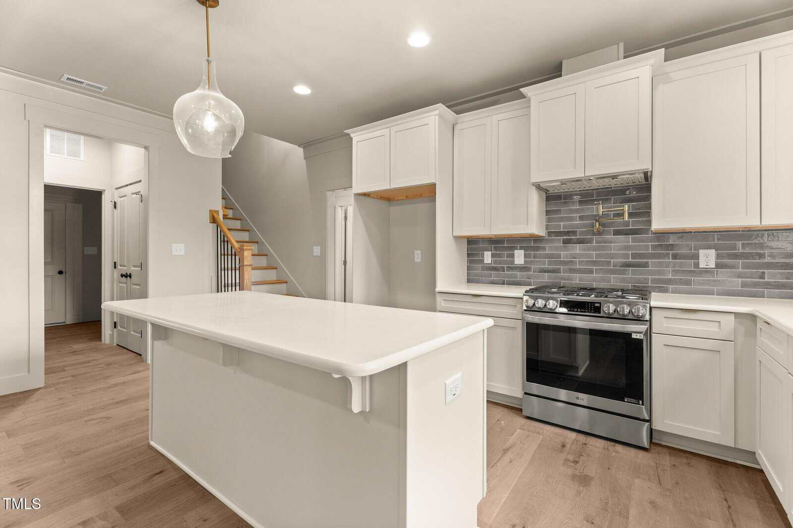 Modern kitchen with white cabinets, gray subway tile backsplash, stainless steel oven, large white island with built-in outlets, pendant lighting, light wood floors, and a staircase in the background.