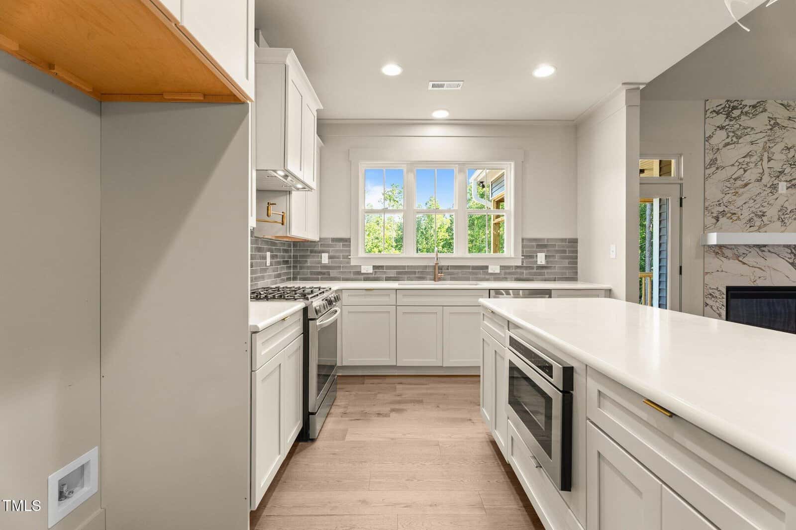 Modern kitchen with white cabinets, gray subway tile backsplash, stainless steel appliances, a built-in microwave, and a large island. Bright window above the sink, wood flooring, and light-colored countertops.