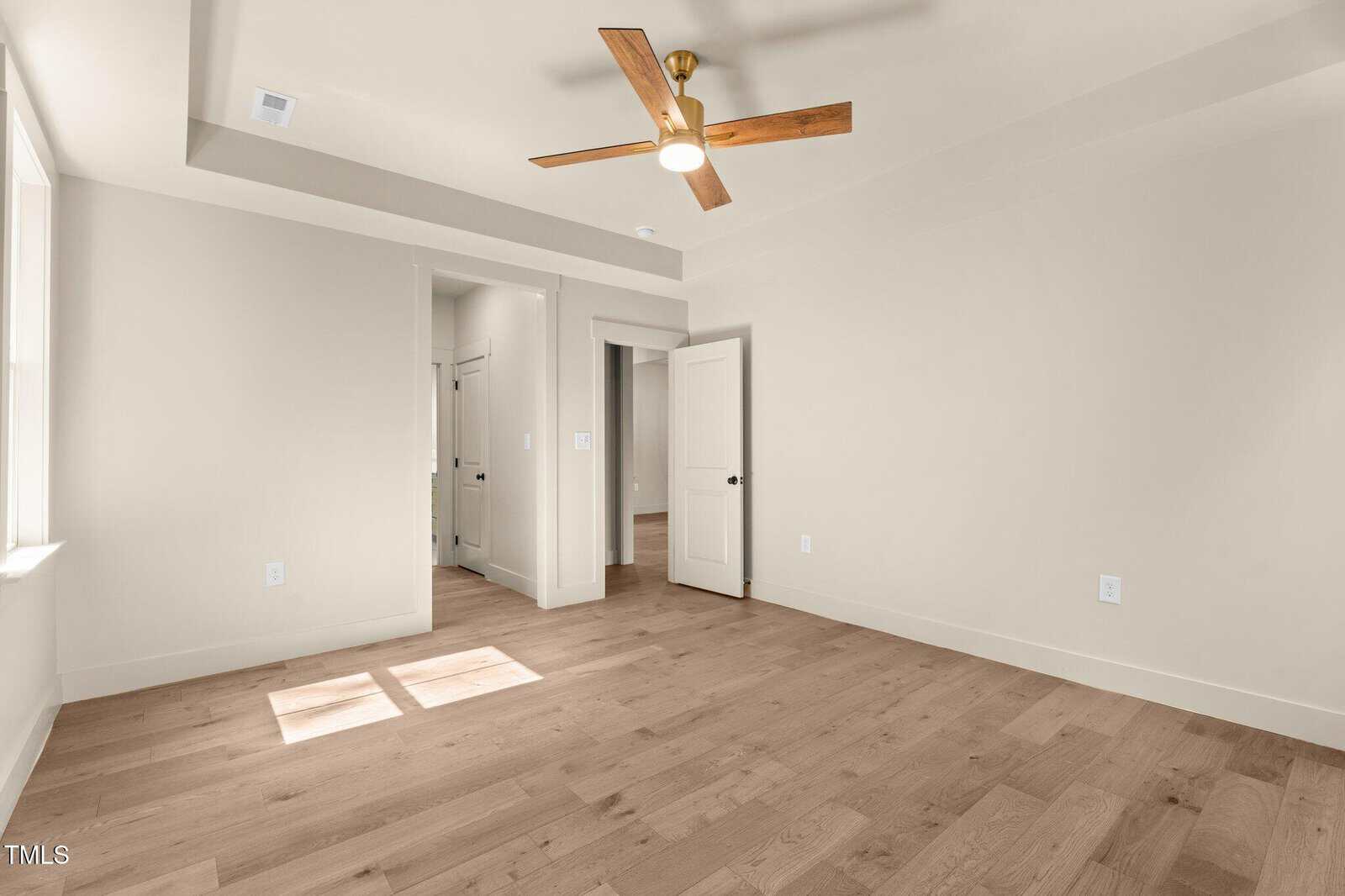 A bright, empty room with light wood flooring, beige walls, a tray ceiling, modern ceiling fan, open door, closet, and natural light streaming through a window.