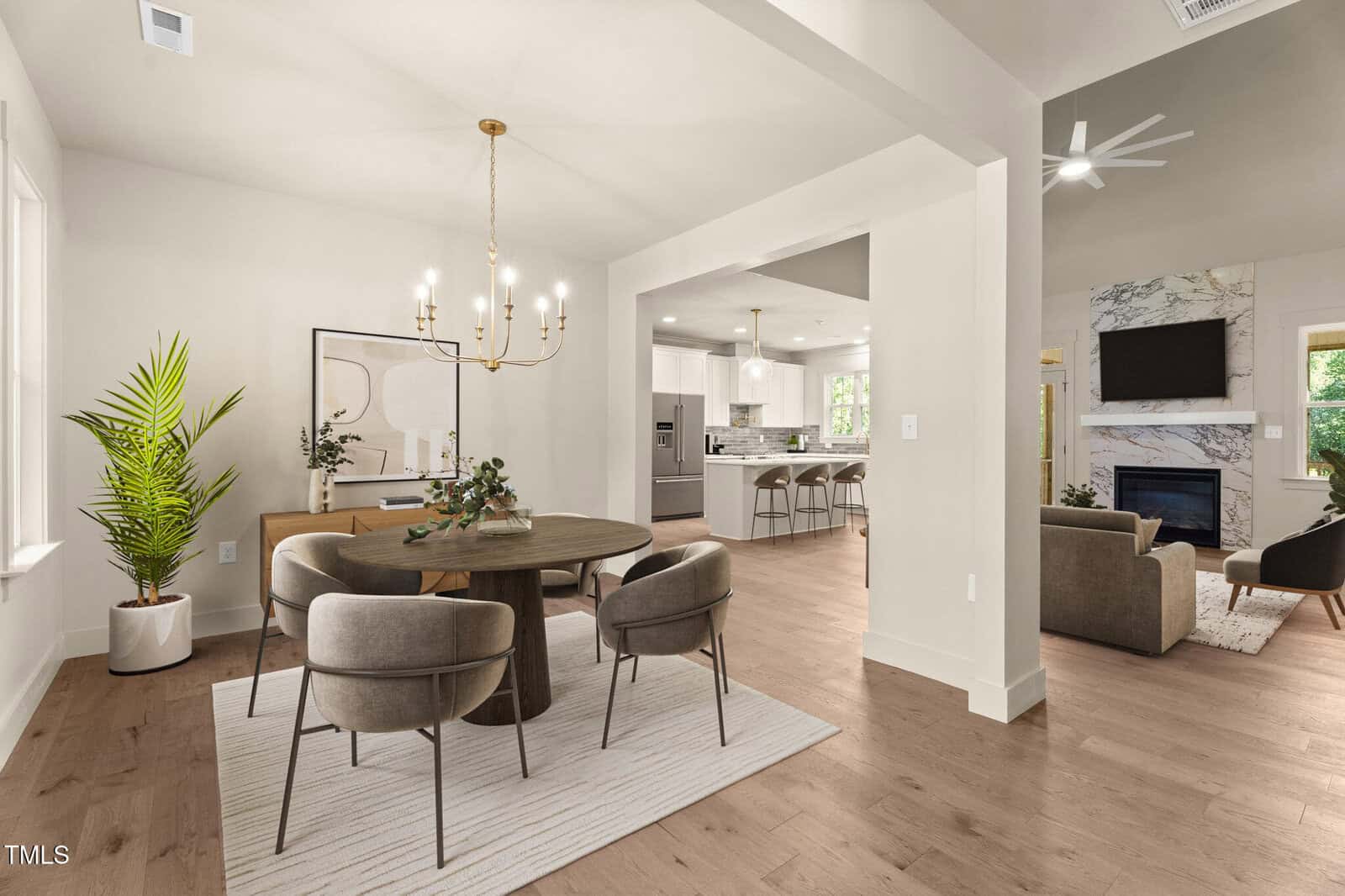 Open-concept living space with a modern dining area featuring a round table and four chairs, a gold chandelier, a potted plant, and view into a bright kitchen and living room with fireplace and TV.