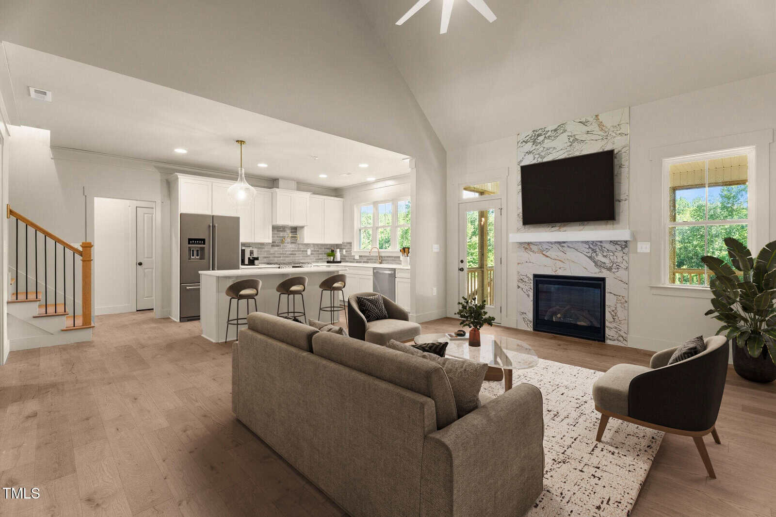 Modern open-concept living room and kitchen with neutral decor, gray sofa set, light wood floors, large windows, marble fireplace, and a kitchen island with bar stools. Staircase visible to the left.