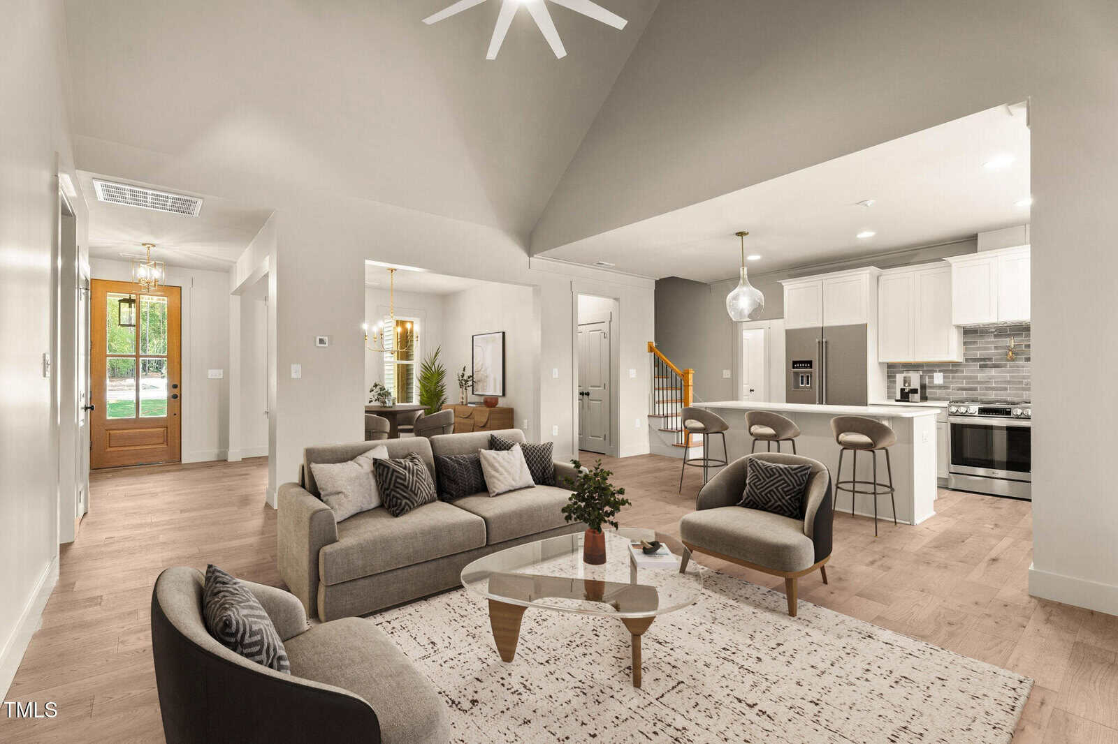 Modern open-concept living room with gray sofas, round coffee table, kitchen island with barstools, dining area, wooden floors, and large windows. Neutral colors and pendant lighting create a cozy, inviting space.