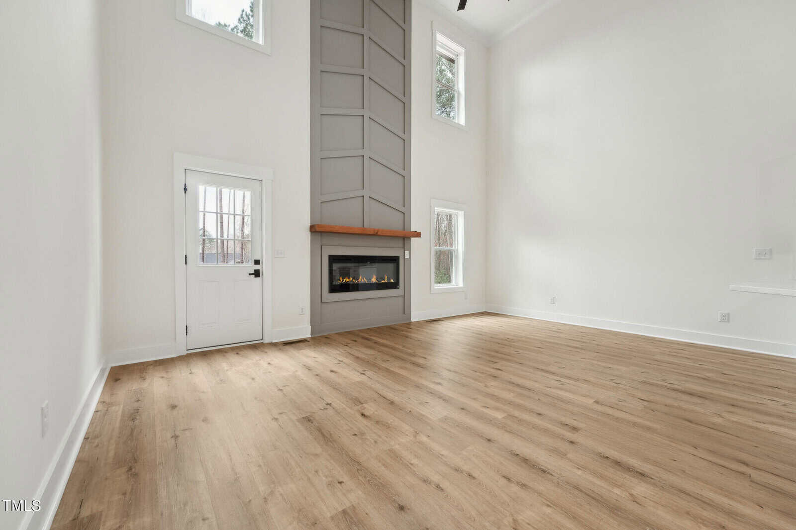 A spacious, empty living room with light wood floors, tall white walls, a gray accent wall with a modern fireplace, multiple windows, and a white door with glass panels.