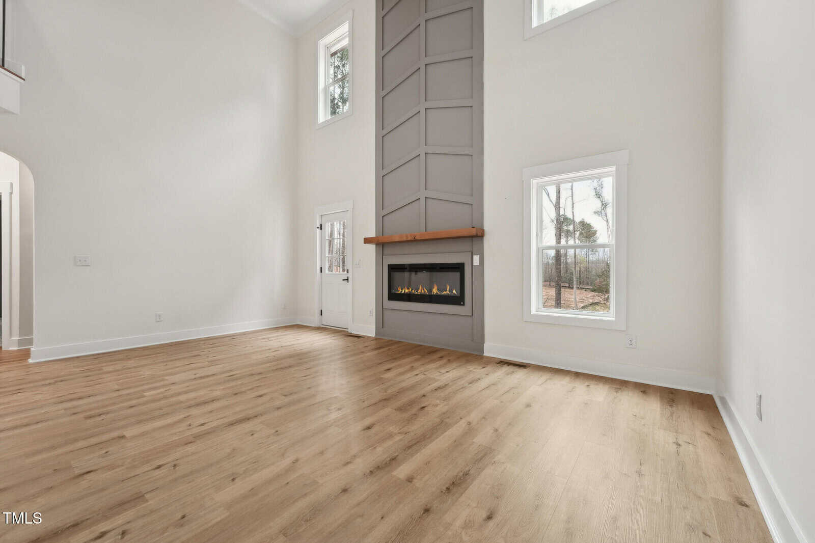 Spacious, modern living room with light wood floors, tall ceilings, white walls, large windows, and a built-in vertical gas fireplace with a gray accent wall and wooden mantel. Natural light fills the room.