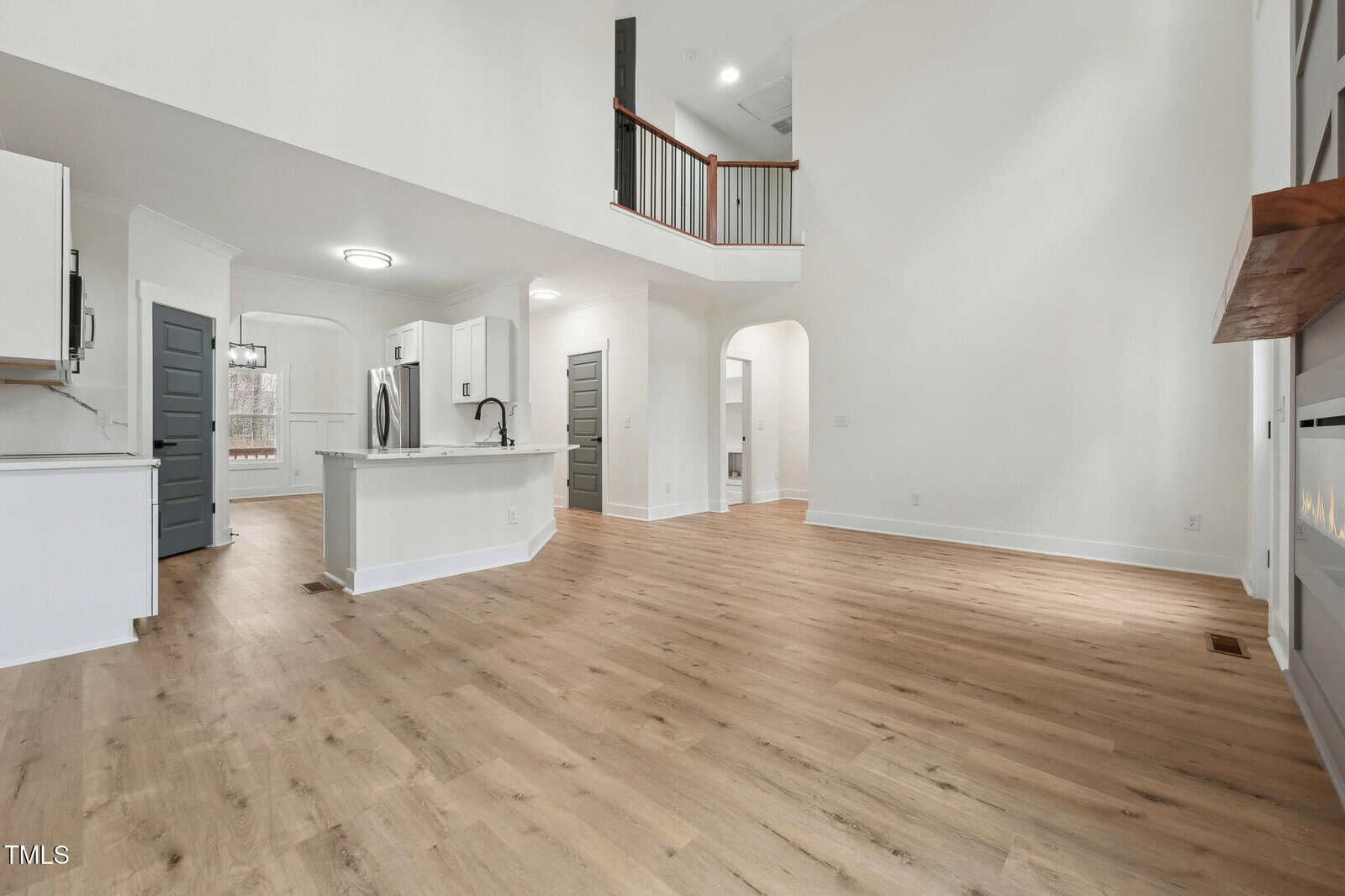 Open-concept living area with light wood floors, white walls, a modern kitchen with stainless steel appliances and island, a staircase with a railing, and a high ceiling with visible upper level.