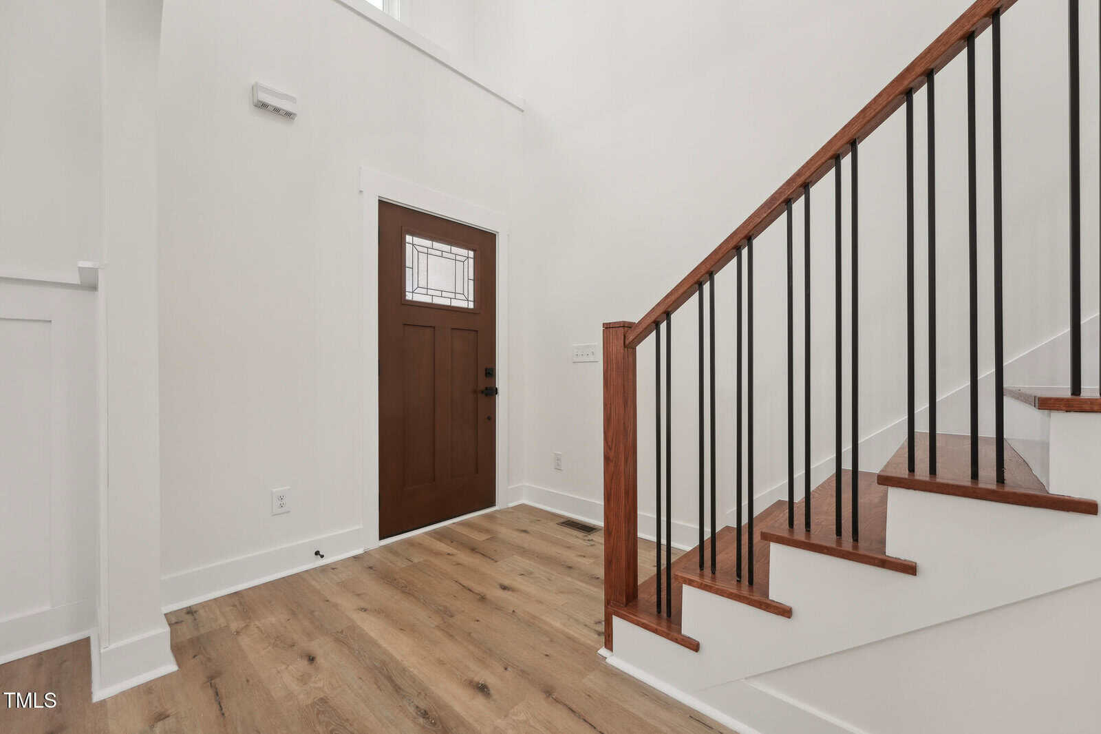 A front entryway with a brown wooden door featuring a glass panel, light wood flooring, white walls, and a staircase with wooden steps and black metal railings.