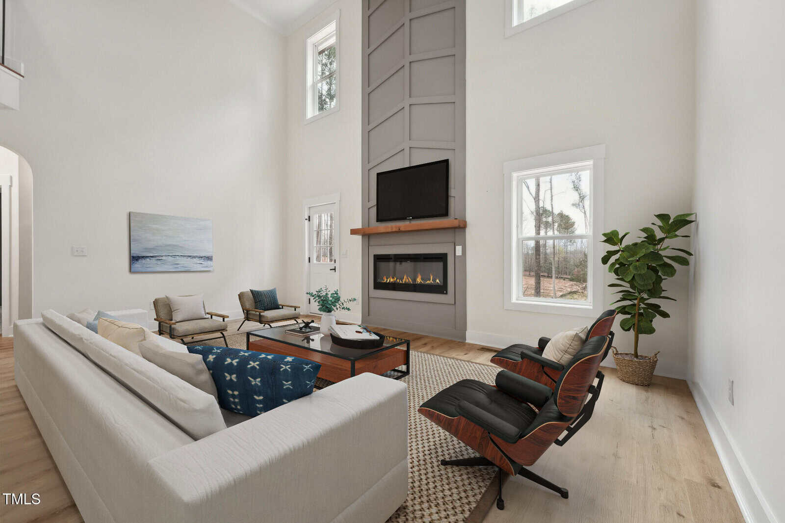 A modern living room with high ceilings, neutral walls, a white sofa, black lounge chair, fireplace with a TV above it, large windows, and a potted plant in the corner.