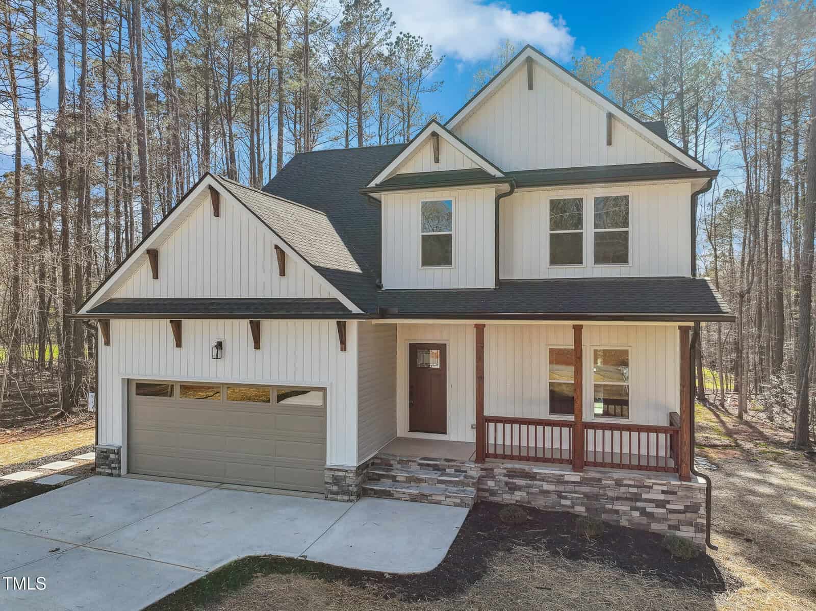 Two-story modern farmhouse with white vertical siding, dark trim, stone accents, a covered front porch with wooden posts, double garage, and concrete driveway set against a background of tall trees.