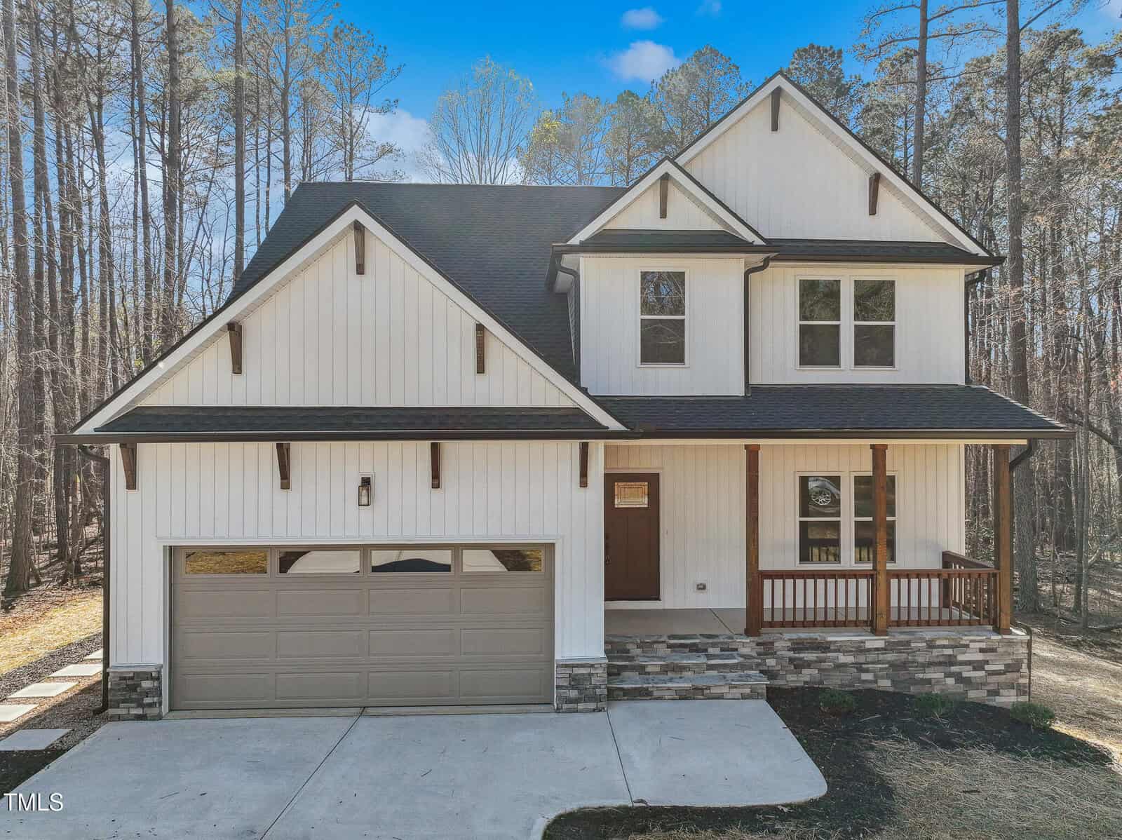 Two-story modern farmhouse with white vertical siding, black trim, a double garage, front porch with wood railing, stone accents on the lower facade, and surrounded by tall trees under a blue sky.